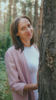 Ağacın arkasından dışarı bakan ciddi bir kadın. Sakin bir kadın ağaca dokunuyor ve sessiz ormanda kameraya bakıyor.