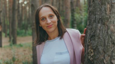 Ağacın arkasından dışarı bakan ciddi bir kadın. Sakin bir kadın ağaca dokunuyor ve sessiz ormanda kameraya bakıyor.