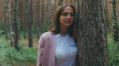 Ağacın arkasından dışarı bakan ciddi bir kadın. Sakin bir kadın ağaca dokunuyor ve sessiz ormanda kameraya bakıyor.