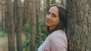 Ağacın arkasından dışarı bakan ciddi bir kadın. Sakin bir kadın ağaca dokunuyor ve sessiz ormanda kameraya bakıyor.