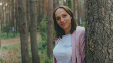 Ağacın arkasından dışarı bakan ciddi bir kadın. Sakin bir kadın ağaca dokunuyor ve sessiz ormanda kameraya bakıyor.