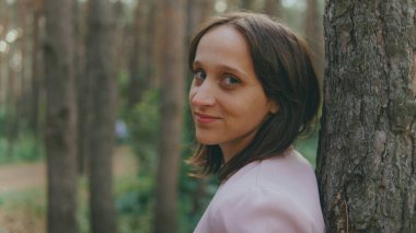 Ağacın arkasından dışarı bakan ciddi bir kadın. Sakin bir kadın ağaca dokunuyor ve sessiz ormanda kameraya bakıyor.
