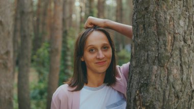 Ağacın arkasından dışarı bakan ciddi bir kadın. Sakin bir kadın ağaca dokunuyor ve sessiz ormanda kameraya bakıyor.
