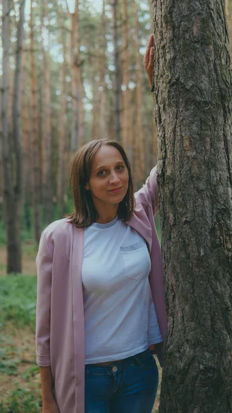 Ağacın arkasından dışarı bakan ciddi bir kadın. Sakin bir kadın ağaca dokunuyor ve sessiz ormanda kameraya bakıyor.