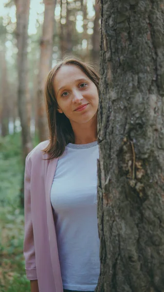 Uma mulher séria a olhar por detrás da árvore. Mulher muito calma tocando árvore e olhando para a câmera na floresta tranquila — Fotografia de Stock