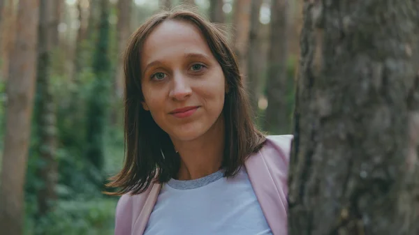 Ernsthafte Frau schaut hinter Baum hervor. Ziemlich ruhige Frau berührt Baum und blickt in ruhigen Wald in die Kamera — Stockfoto