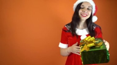 Noel Baba kostümüyle gülümseyen genç bir kadın turuncu arka planda hediyeler tutuyor. Mutlu Noeller ve mutlu yıllar..