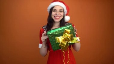 Noel Baba kostümüyle gülümseyen genç bir kadın turuncu arka planda hediyeler tutuyor. Mutlu Noeller ve mutlu yıllar..