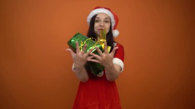 Noel Baba kostümüyle gülümseyen genç bir kadın turuncu arka planda hediyeler tutuyor. Mutlu Noeller ve mutlu yıllar..