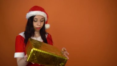 Noel Baba kostümüyle gülümseyen genç bir kadın turuncu arka planda hediyeler tutuyor. Mutlu Noeller ve mutlu yıllar..