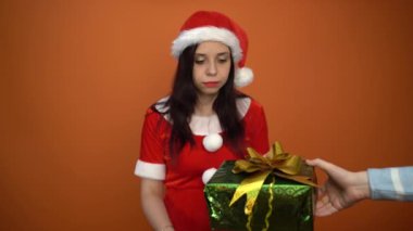 Noel Baba kostümüyle gülümseyen genç bir kadın turuncu arka planda hediyelerle dolu bir kutu tutuyor. Mutlu Noeller ve mutlu yıllar..