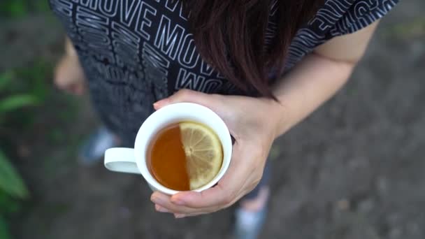Девочка держит в руках чашку чая с лемоном. Женщина в прохладном летнем чае пьет чай на улице. — стоковое видео