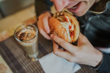 Genç adam hamburger yiyor. Bir adam masada oturuyor ve fast food kafede çizburger yiyor..