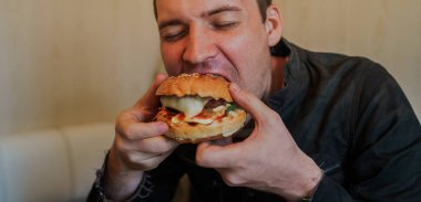 Genç adam hamburger yiyor. Bir adam masada oturuyor ve fast food kafede çizburger yiyor..