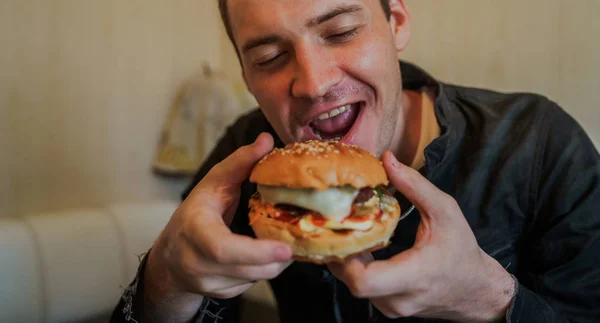 Bonjour, je mange de la nourriture rapide, du hamburger. Un homme est assis à la table et mange un hamburger au fromage — Photo