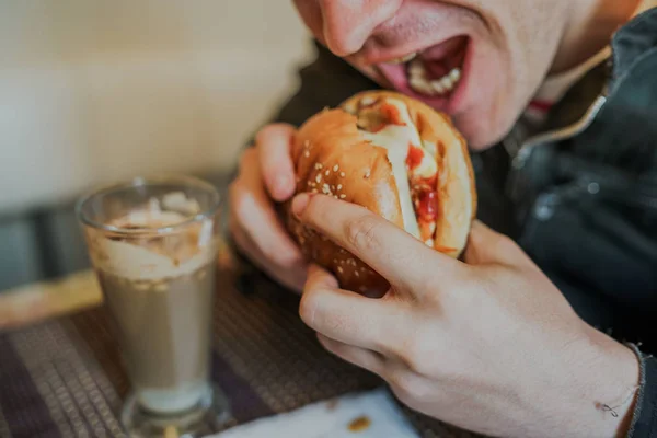 Mladý muž jí hamburger. Muž sedí u stolu a jí cheeseburger v kavárně s rychlým občerstvením. — Stock fotografie