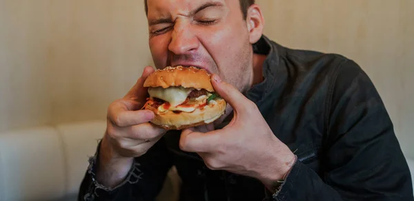 El joven está comiendo hamburguesas. Un hombre está sentado a la mesa y comiendo un queseburger en un café de comida rápida.. — Foto de Stock