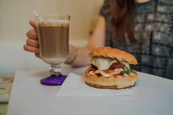 Food-Fotografie. Heißer Cappuccino mit Hamburger am Tisch im Fast-Food-Café. Cheeseburger und Kaffee auf dem Tisch, Getränk enthält Koffein. — Stockfoto