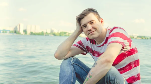 Man by the river.Portrait of a male in a t-shirt sitting on a column near the river on a Sunny day — Stock Photo, Image