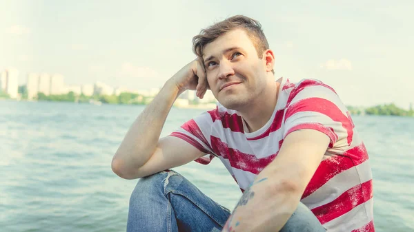 Man bij de rivier.Portret van een mannetje in een t-shirt zittend op een zuil bij de rivier op een zonnige dag — Stockfoto