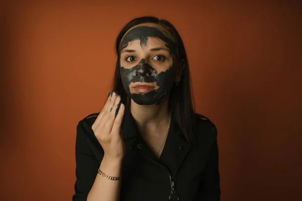 Una joven inflige una máscara negra en la cara sobre un fondo naranja. El concepto de estilo de vida saludable, belleza, cuidado corporal.. — Foto de Stock