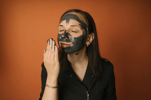 Молода жінка наносить чорну маску на обличчя на помаранчевий фон. Концепція здорового способу життя, краси, догляду за тілом . — стокове фото