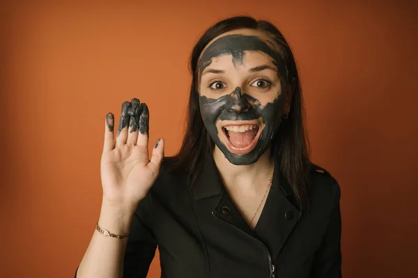 Eine junge Frau setzt dem Gesicht auf orangefarbenem Hintergrund eine schwarze Maske auf. Das Konzept der gesunden Lebensweise, Schönheit, Körperpflege. — Stockfoto