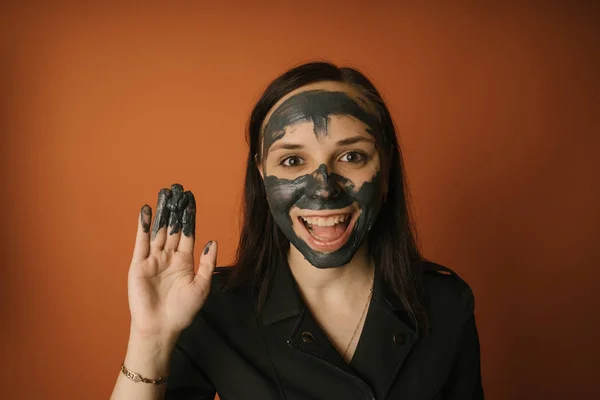 Genç bir kadın turuncu arka planda yüzüne siyah bir maske takıyor. Sağlıklı yaşam tarzı, güzellik, vücut bakımı kavramı. — Stok fotoğraf