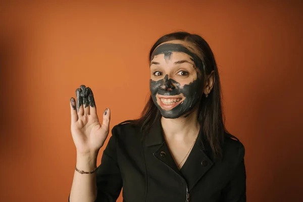 Genç bir kadın turuncu arka planda yüzüne siyah bir maske takıyor. Sağlıklı yaşam tarzı, güzellik, vücut bakımı kavramı. — Stok fotoğraf