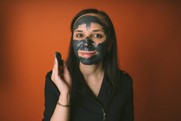 A young woman inflicts a black mask on the face on an orange background. The concept of healthy lifestyle, beauty, body care.