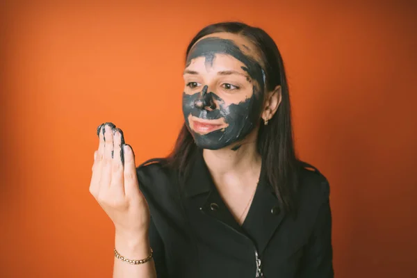 Une jeune femme pose un masque noir sur le visage sur fond orange. Le concept de mode de vie sain, de beauté et de soins corporels. — Photo