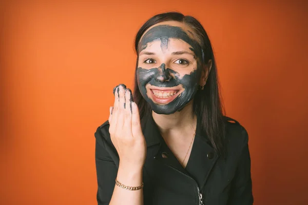 En ung kvinna lägger en svart mask i ansiktet på en orange bakgrund. Begreppet hälsosam livsstil, skönhet, kroppsvård. — Stockfoto