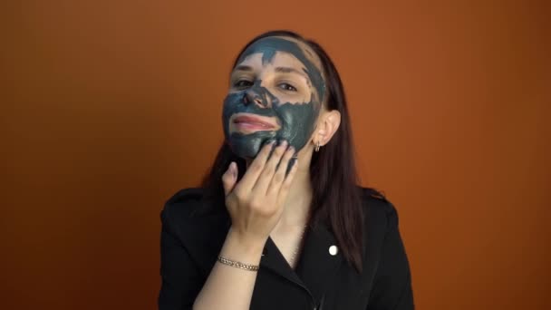 En ung kvinna lägger en svart mask i ansiktet på en orange bakgrund. Begreppet hälsosam livsstil, skönhet, kroppsvård. — Stockvideo