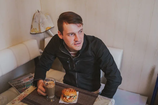 Un homme prend le petit déjeuner, assis à la table. Café et hamburger sur la table . — Photo