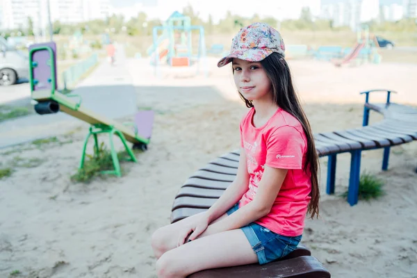 O fată cu părul lung și întunecat în haine casual stă pe o bancă pe terenul de joacă . — Fotografie, imagine de stoc