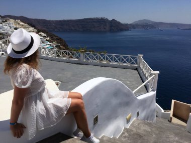 Beyaz elbiseli ve beyaz şapkalı genç bir kadın denize bakıyor. Oia, Santorini, Yunanistan adasında tatilde olan bir turist kadın. Genç bir kadın ünlü beyaz köyü ziyaret ediyor.