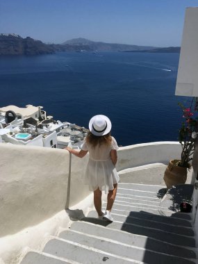 Beyaz elbiseli ve beyaz şapkalı genç bir kadın denize bakıyor. Oia, Santorini, Yunanistan adasında tatilde olan bir turist kadın. Genç bir kadın ünlü beyaz köyü ziyaret ediyor.