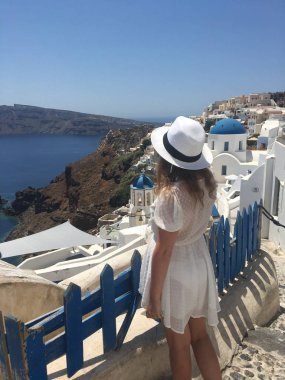 Beyaz elbiseli ve beyaz şapkalı genç bir kadın denize bakıyor. Oia, Santorini, Yunanistan adasında tatilde olan bir turist kadın. Genç bir kadın ünlü beyaz köyü ziyaret ediyor.