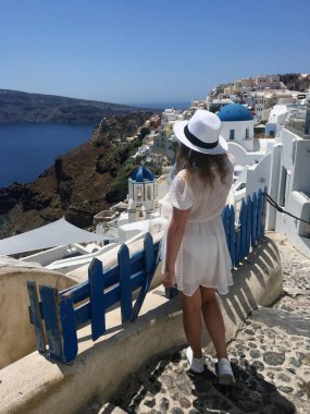 Beyaz elbiseli ve beyaz şapkalı genç bir kadın denize bakıyor. Oia, Santorini, Yunanistan adasında tatilde olan bir turist kadın. Genç bir kadın ünlü beyaz köyü ziyaret ediyor.