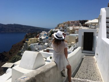 Beyaz elbiseli ve beyaz şapkalı genç bir kadın denize bakıyor. Oia, Santorini, Yunanistan adasında tatilde olan bir turist kadın. Genç bir kadın ünlü beyaz köyü ziyaret ediyor.