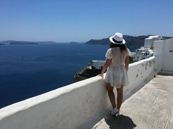 Una joven vestida de blanco y sombrero blanco mira al mar. Mujer de turismo de vacaciones en Oia, Santorini, isla de Grecia. Una joven está visitando el famoso pueblo blanco con el — Foto de Stock