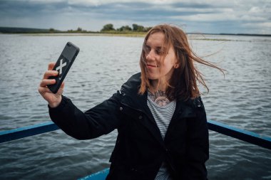 Genç bir kadın bulutlu bir havada arka planda cep telefonuyla selfie çekiyor..