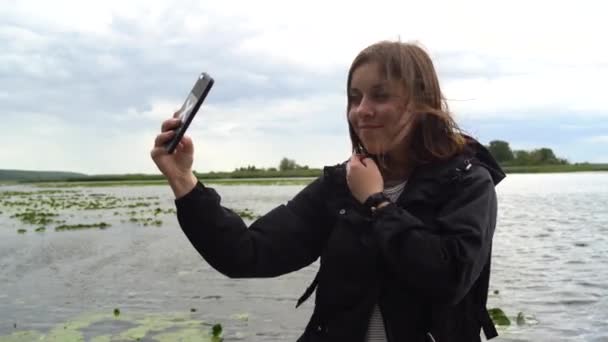 Een jonge vrouw neemt selfie op mobiele telefoon op de achtergrond van water bij bewolkt weer. — Stockvideo