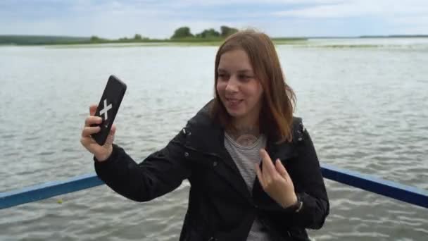 Eine junge Frau macht bei trübem Wetter ein Selfie auf dem Handy vor dem Hintergrund des Wassers. — Stockvideo