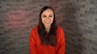 Güzel, genç bir esmer havasında. Kırmızı kapüşonlu güzel bir kadın mutlu bir şekilde güler ve tuğla bir arka planda olumlu duygular ifade eder..