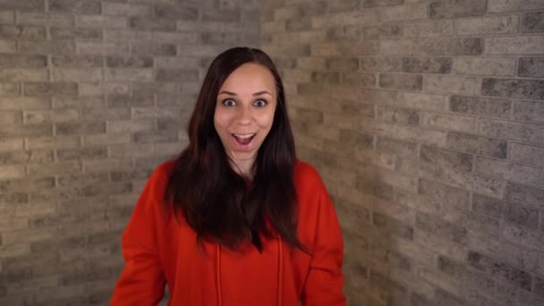 Uma mulher emocional bonita em um hoodie vermelho é surpreendida feliz com as mãos em suas bochechas. — Vídeo de Stock