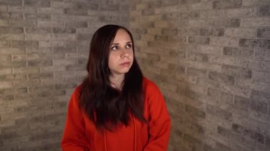 Kırmızı kapüşonlu güzel, duygusal bir kadın şüpheli bir şekilde tuğla arka planda etrafına bakıyor..