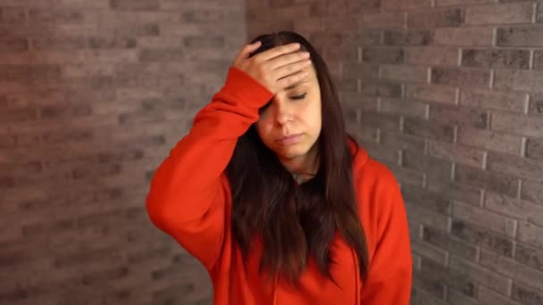 A beautiful emotional woman in a red hoodie is doing facepalm covering her face with palm. A young woman in a state of shock is covering her face with palm. – Stock-video