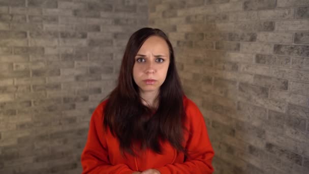 A beautiful emotional woman in a red hoodie raises her eyebrows in surprise on a brick background. — Stock Video