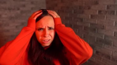 Kırmızı kapüşonlu genç bir kadın ellerini tuğla bir arka planda acı içinde tutuyor. Bir kadının başı ağrır..